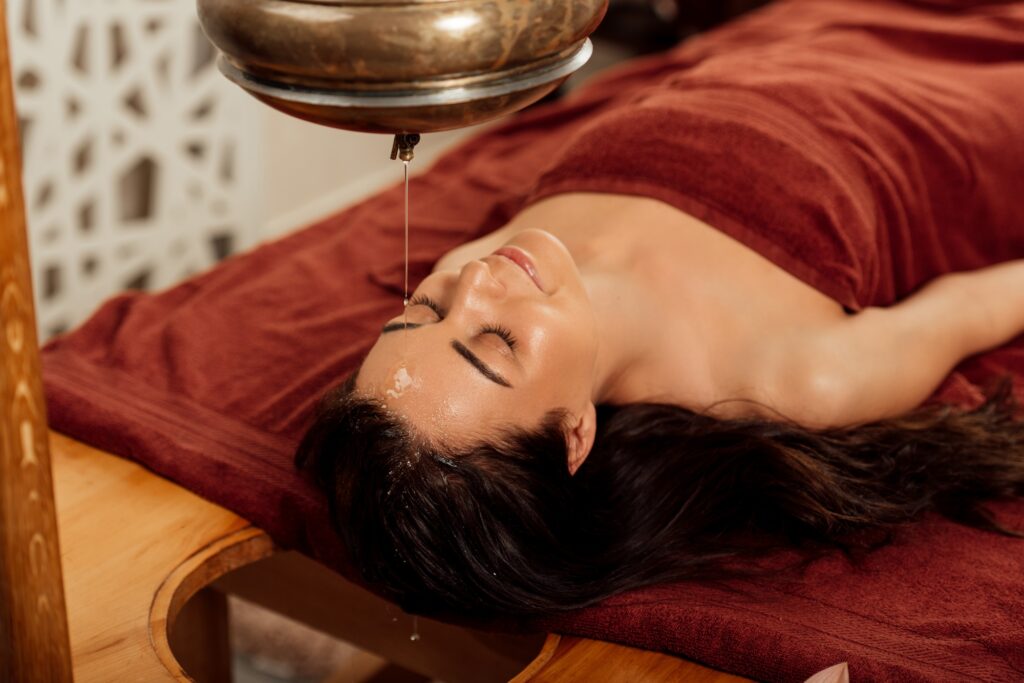 relaxed young woman lying under shirodhara vessel during ayurvedic procedure